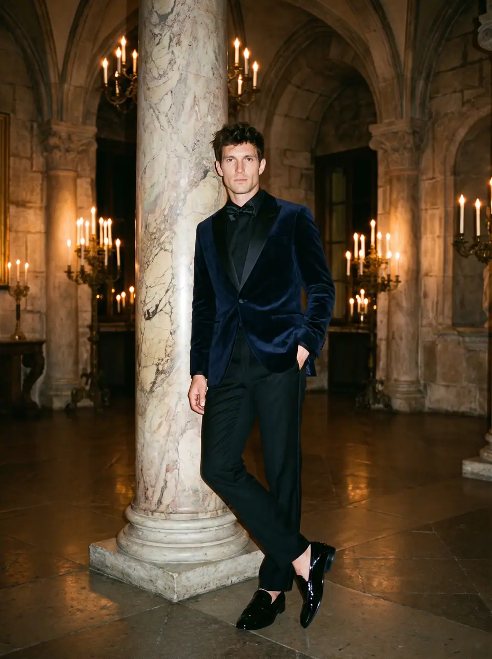 A man in a midnight-blue velvet dinner jacket and black trousers leans on a marble column in a candlelit hall.