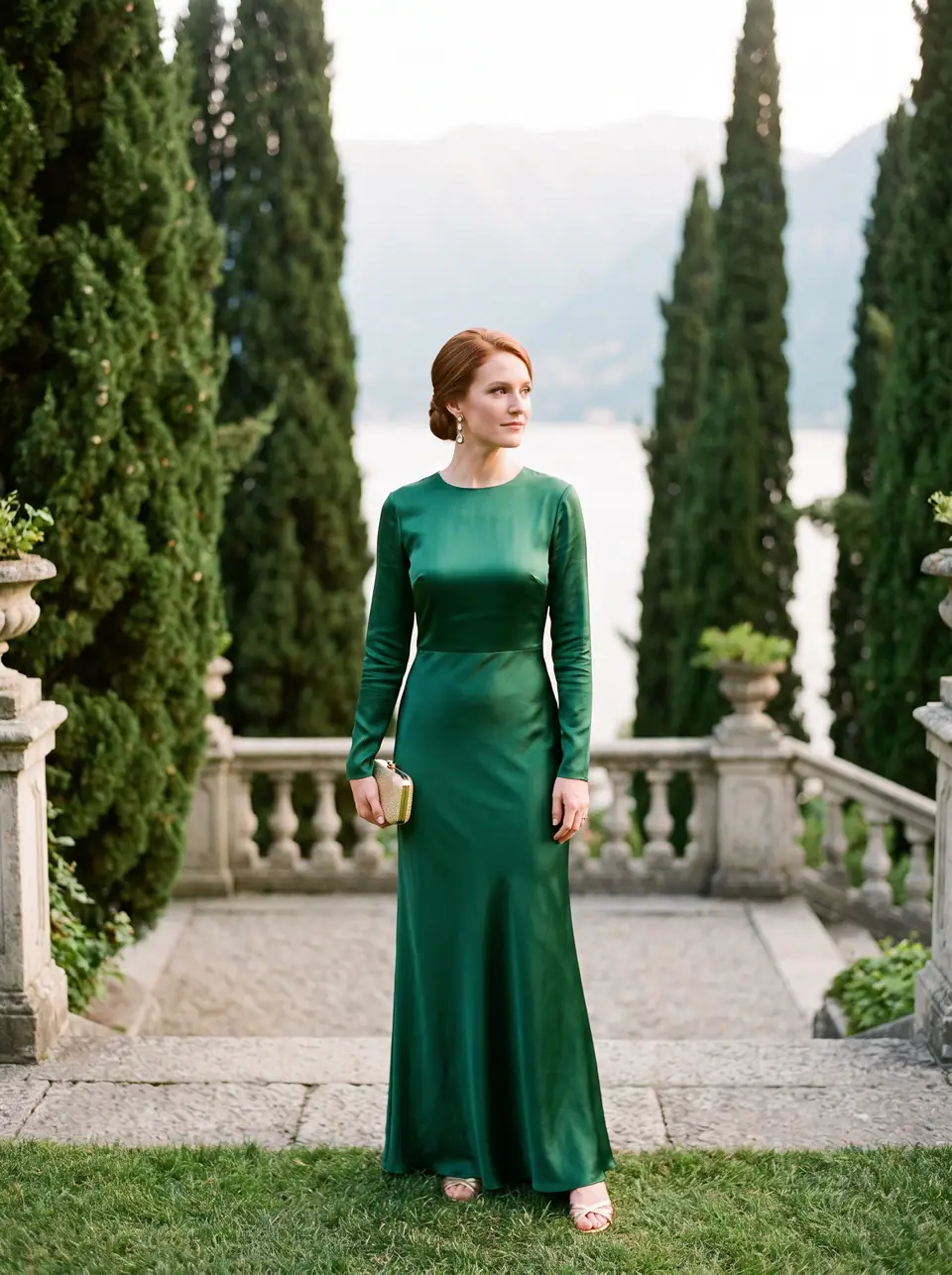 A woman in a long-sleeved emerald satin gown stands on a garden terrace between cypress trees above Lake Como.