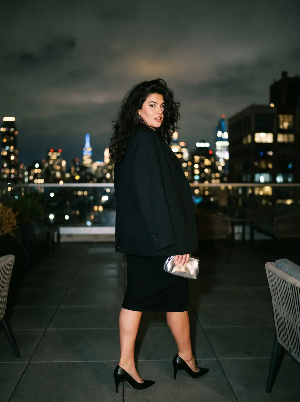 A woman in a black blazer worn over the shoulders and a black pencil skirt looks back on a rooftop at night, the skyline lit behind her.